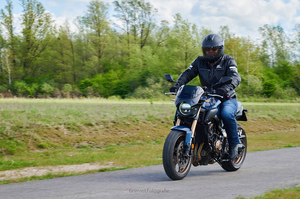 Alt tekst: Elektrische motorfiets rijden door groen landschap met man in helm en lederen jas, scherp en dynamisch, voor een energiezuinig vervoer en avontuurlijke reizen.