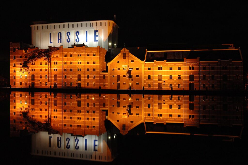 Industrieel gebouw verlicht met de bedrijfsnaam "LASSIE" op een rotsachtig gevel, reflectie in het water, nachtelijke sfeer, moderne en creatieve locatie, branding en marketing concept, ontwerp en beeldvorming.