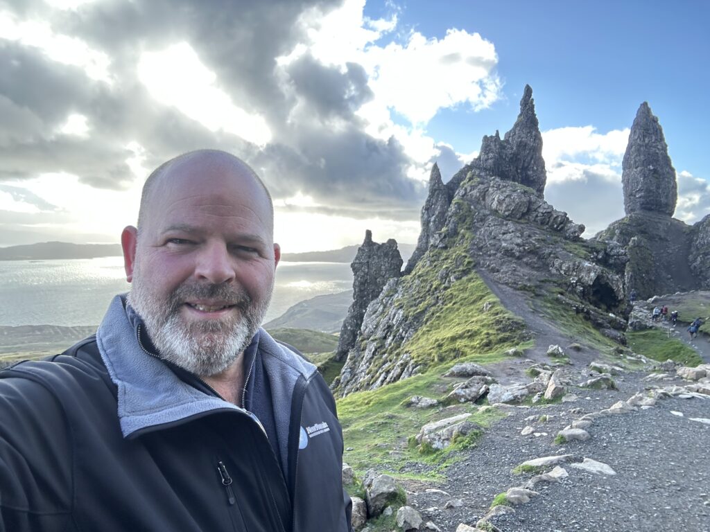 Foto van Frans die poseert voor de iconische rotsformatie op Skye, Schotland, met een uitgestrekt landschap en een dramatische lucht op de achtergrond. Perfect voor natuurlijke avontuurlijke content en reiservaringen.