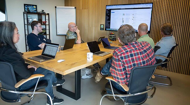 Zes collega's van Sturdy Digital hebben een vergadering in de vergaderruimte en kijken allemaal naar het scherm waarop de presentatie wordt getoond.