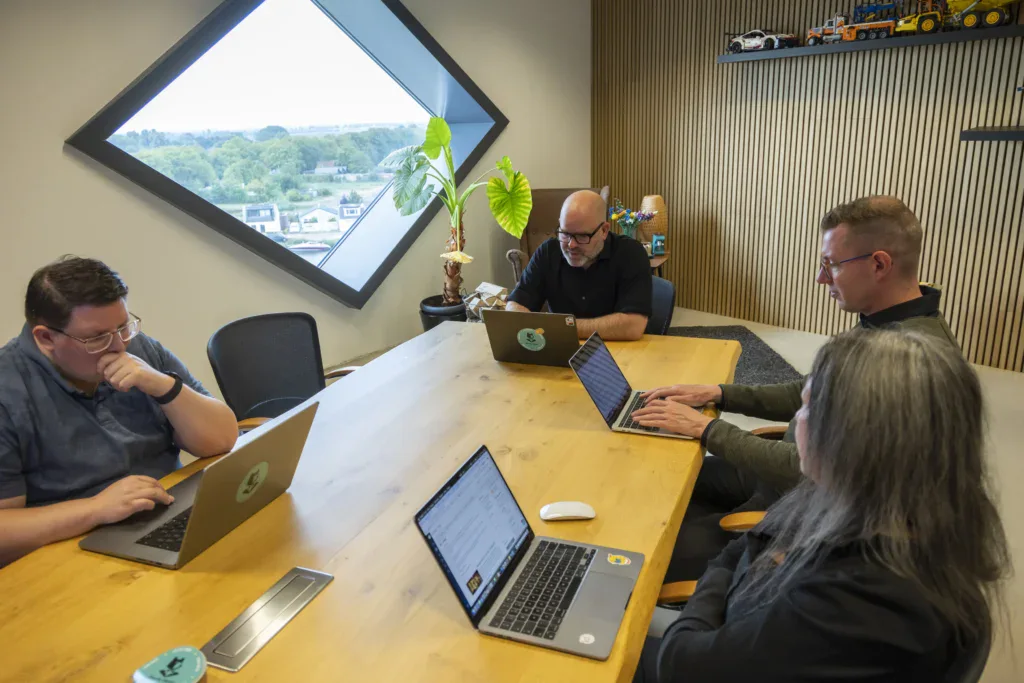 Collega's zitten aan een tafel in een vergaderkamer op kantoor en kijken op hun laptop.