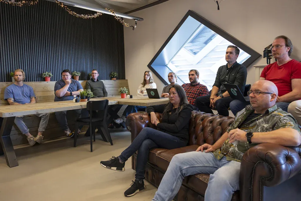 Het team luistert naar een presentatie in de kantine en ze kijken allemaal naar een scherm die buiten het beeld staat.