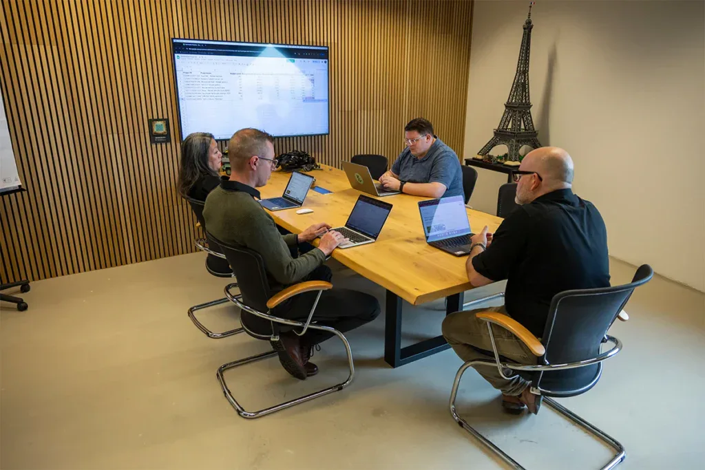 Het team overlegt aan tafel en kijkt naar een groot scherm in de vergaderruimte.