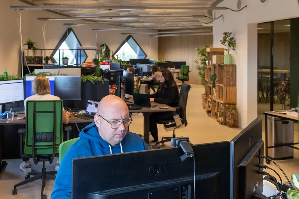 Back end developer programmeert achter zijn bureau. Op de achtergrond zie je collega's aan het werk.