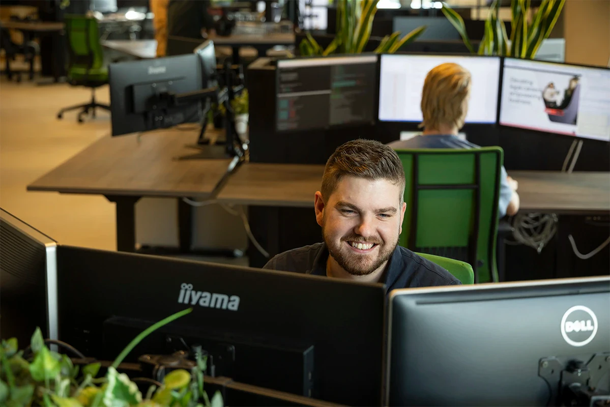 Front end developer zit lachend achter zijn bureau te werken. In de achtergrond zie je collega's aan het werk.