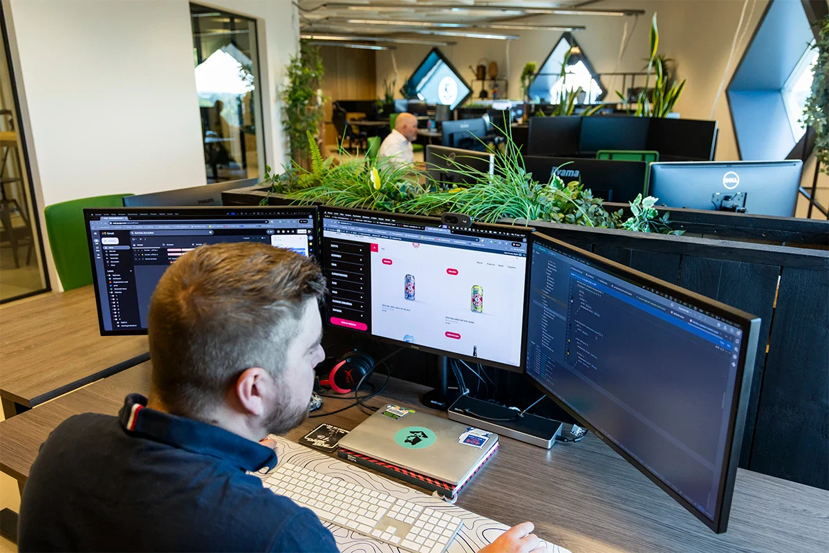 Front end developer programmeert achter zijn bureau op kantoor. In de achtergrond zijn collega's aan het werk.