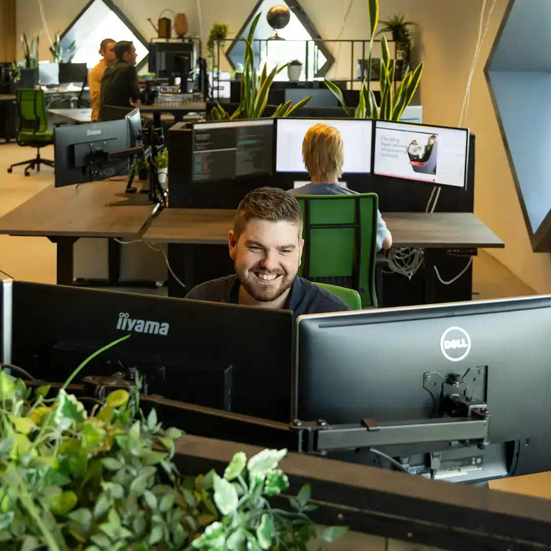 Front end developer zit lachend achter zijn bureau te werken, op kantoor. In de achtergrond zie je collega's die ook werken.