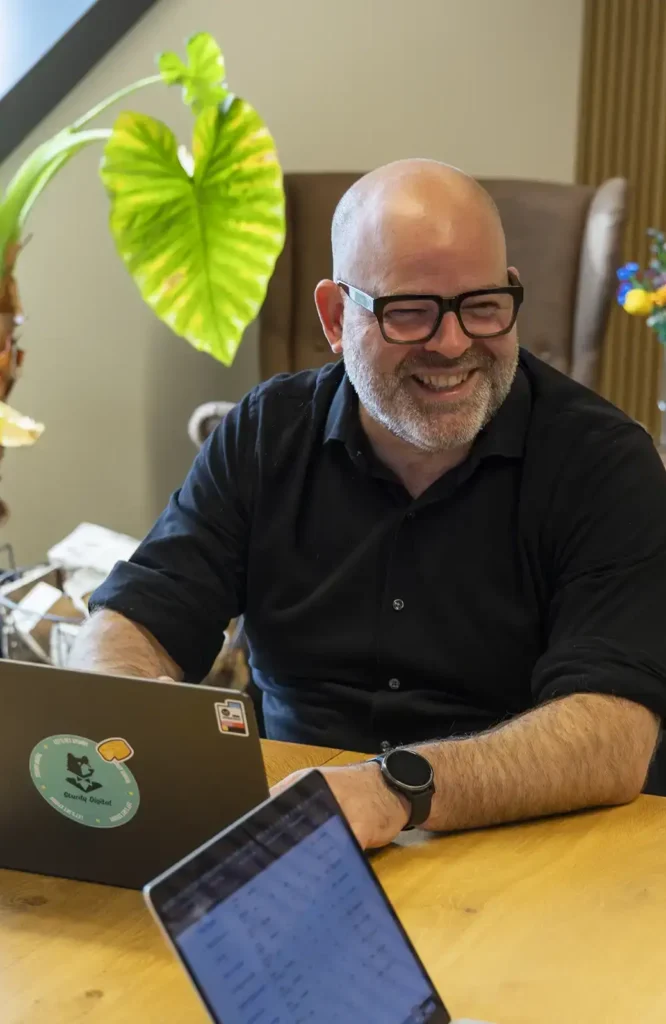 Online marketing expertérieure man die lachend voor laptop zit, met groene plant op de achtergrond.